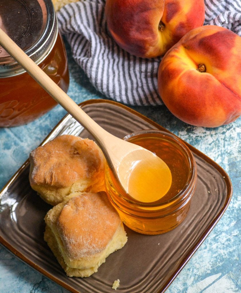 homemade peach syrup in a glass jar on a brown plate next to buttermilk biscuits