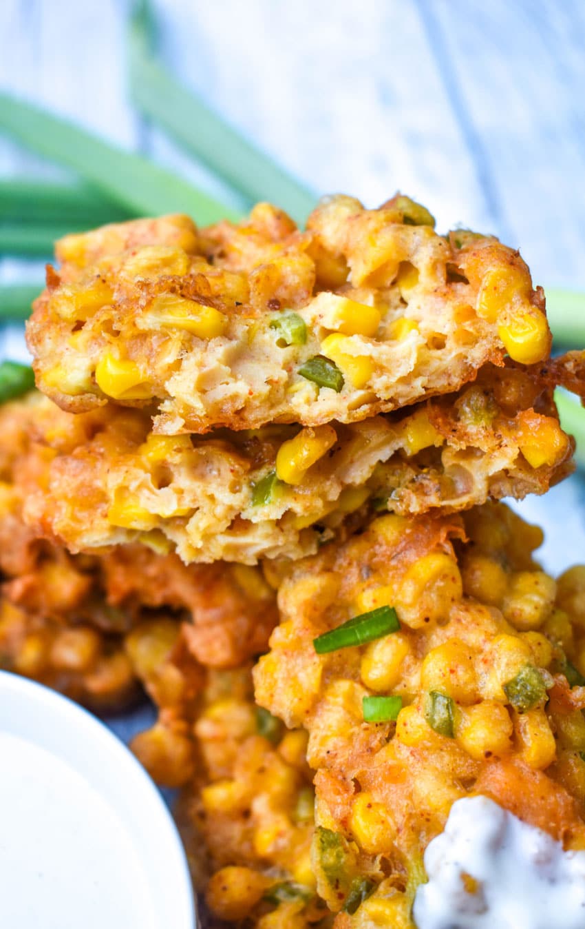 a stack of cheesy corn fritters with the top one broken in half to reveal the center on a wooden table