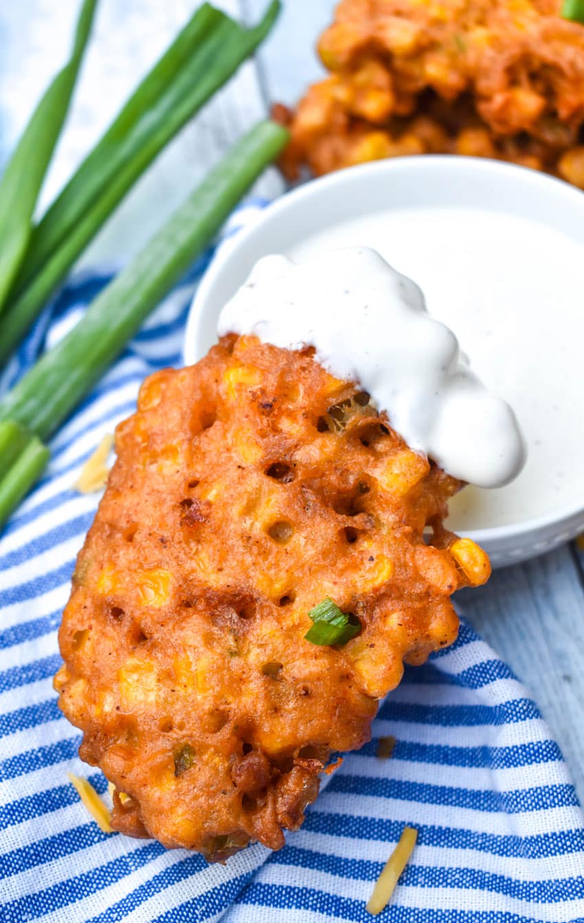 a ranch dipped cheesy corn fritter resting on the edge of a small white bowl filled with ranch dressing