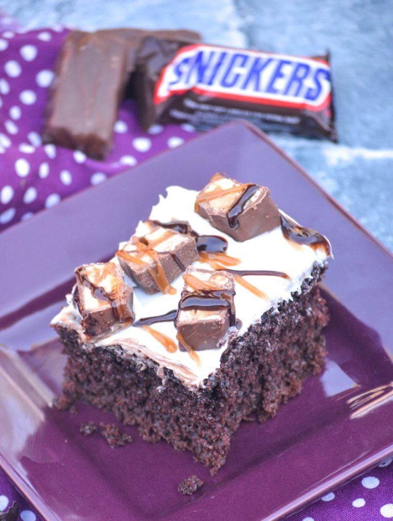 a fluffy piece of vanilla frosting topped snickers poke cake served on a purple appetizer plate