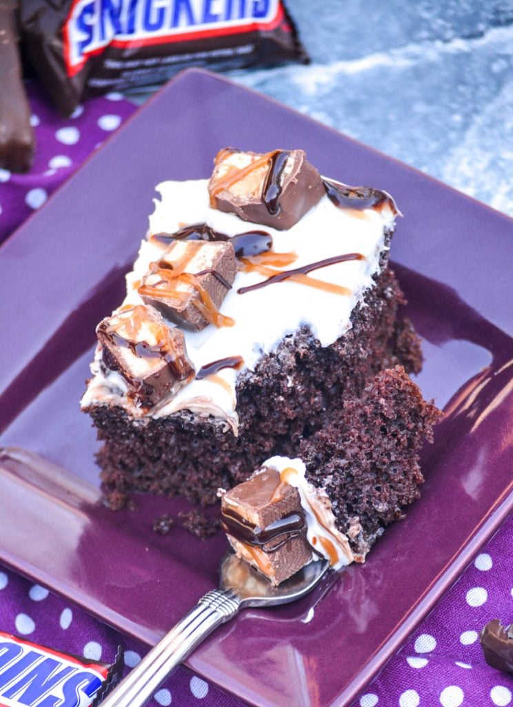 a fluffy piece of vanilla frosting topped snickers poke cake served on a purple appetizer plate with a fork scooping a bite