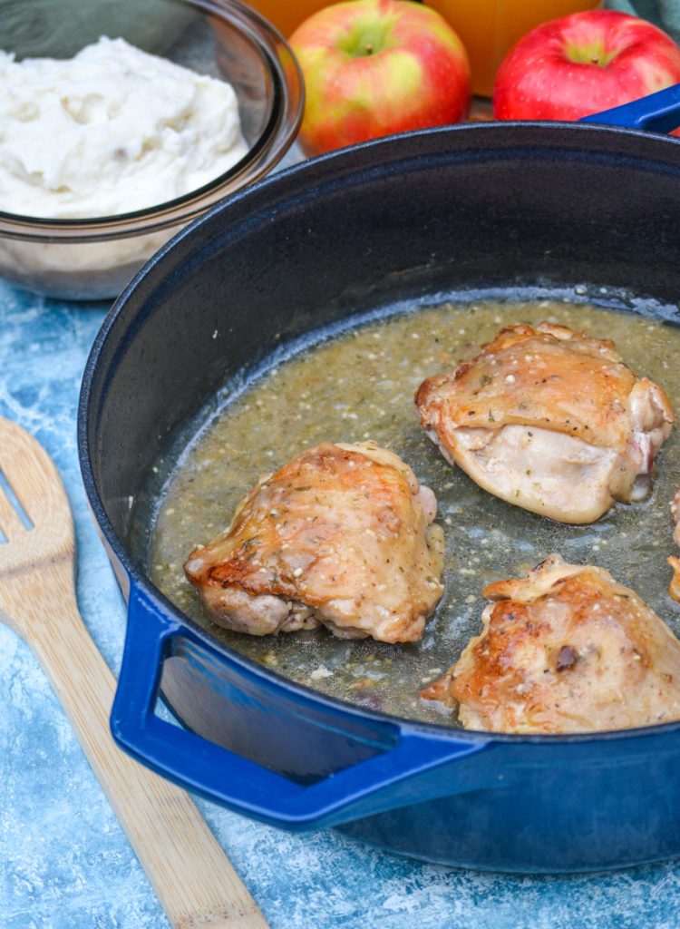 pan fried chicken bathed in a savory apple cider gravy in a blue pot with apples and mashed potatoes in the background and a wooden spoon off to the side for serving
