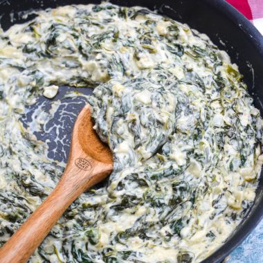 a wooden spoon shown ladling up some of the creamed spinach from black skillet