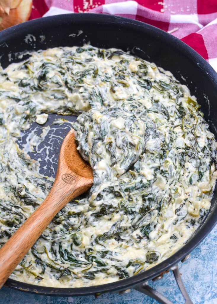 a wooden spoon shown ladling up some of the creamed spinach from black skillet