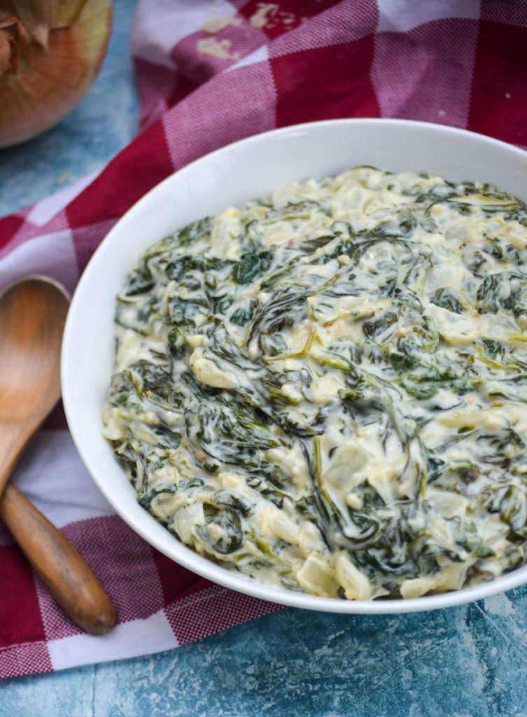 creamed spinach served in a shallow white bowl on a checkered cloth napkin with wooden spoons off to the side