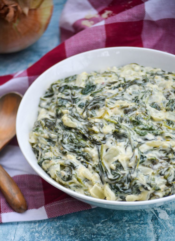 creamed spinach served in a shallow white bowl on a checkered cloth napkin with wooden spoons off to the side