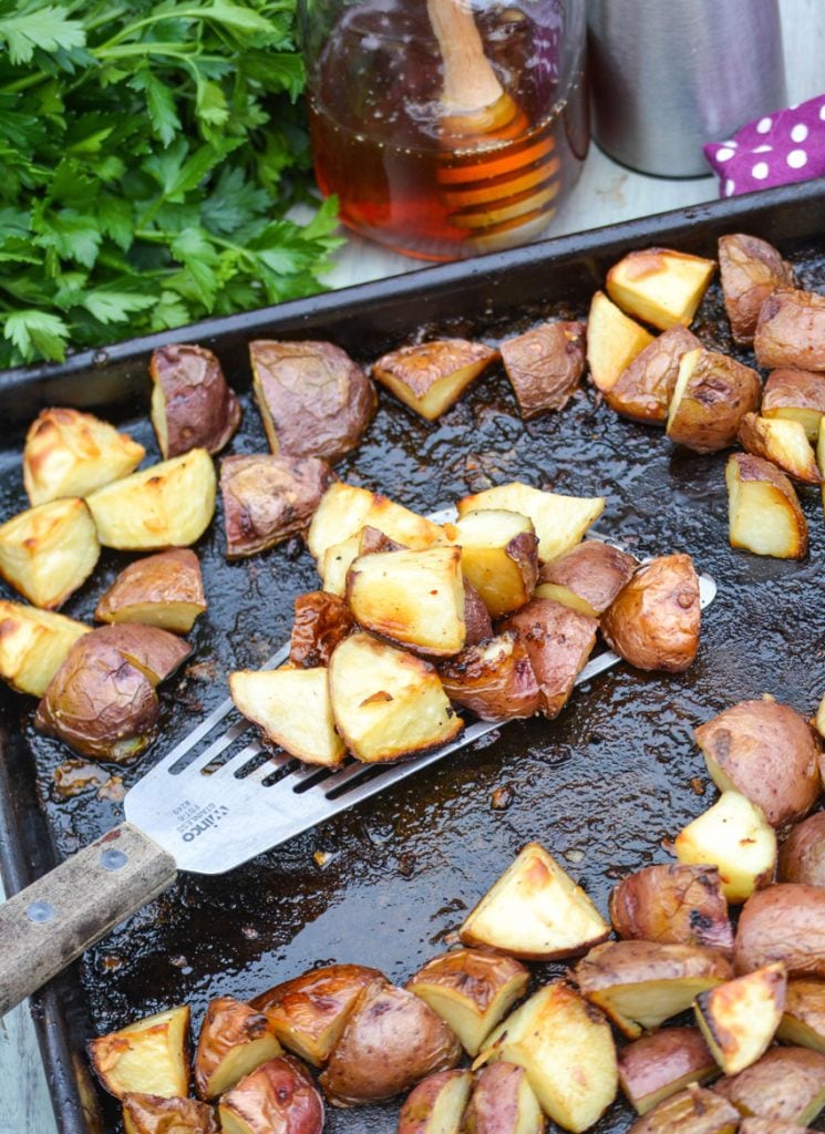 a metal spatula scooping up a serving of honey roasted red potatoes from a dark baking sheet