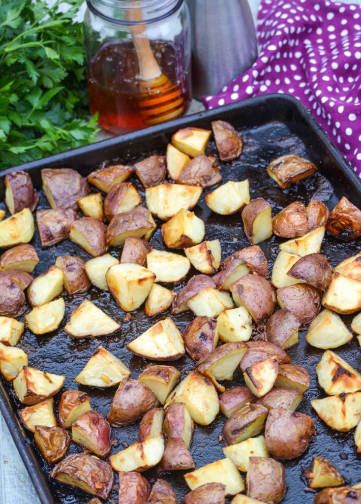 honey roasted red potatoes on a dark brown sheet pan with a brunch of fresh parsley and a jar of honey in the background