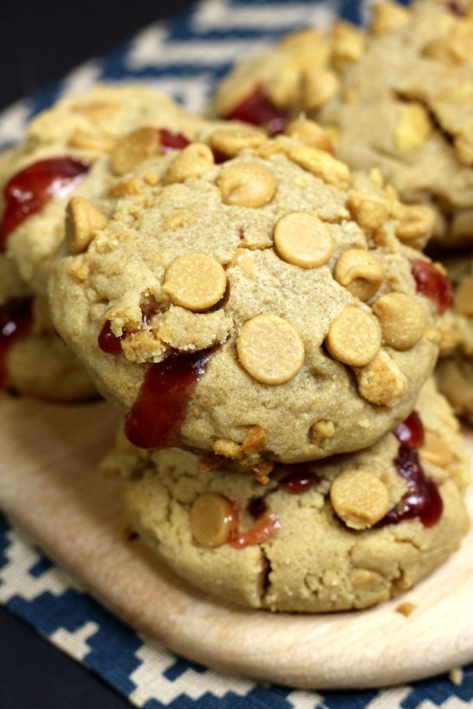 peanut butter jelly cookies are stuffed with strawberry jelly and studded with peanut butter chocolate chips and piled on a wooden cutting board
