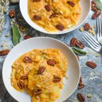 pumpkin alfredo sauce tossed with ravioli served in two white bowls with sage leaves and pecans in the background