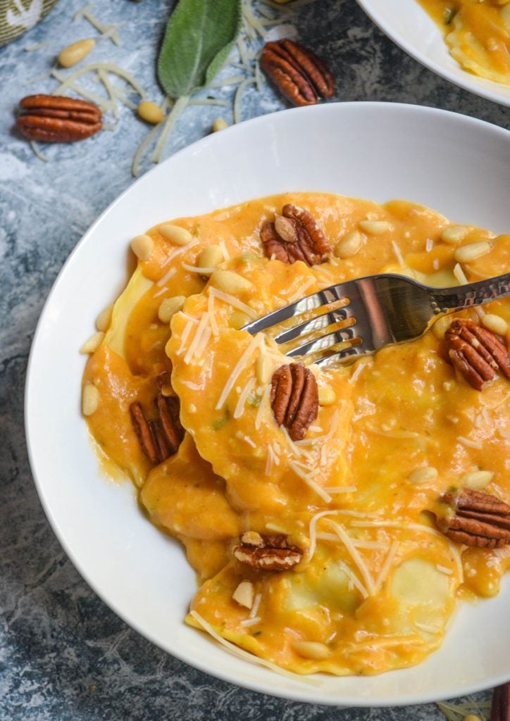 pumpkin alfredo sauce tossed with ravioli served in two white bowls with sage leaves and pecans in the background