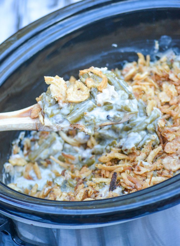 slow cooker green bean casserole topped with crispy fried onions in the bowl of a crockpot with a wooden spoon lifting up a generous scoop