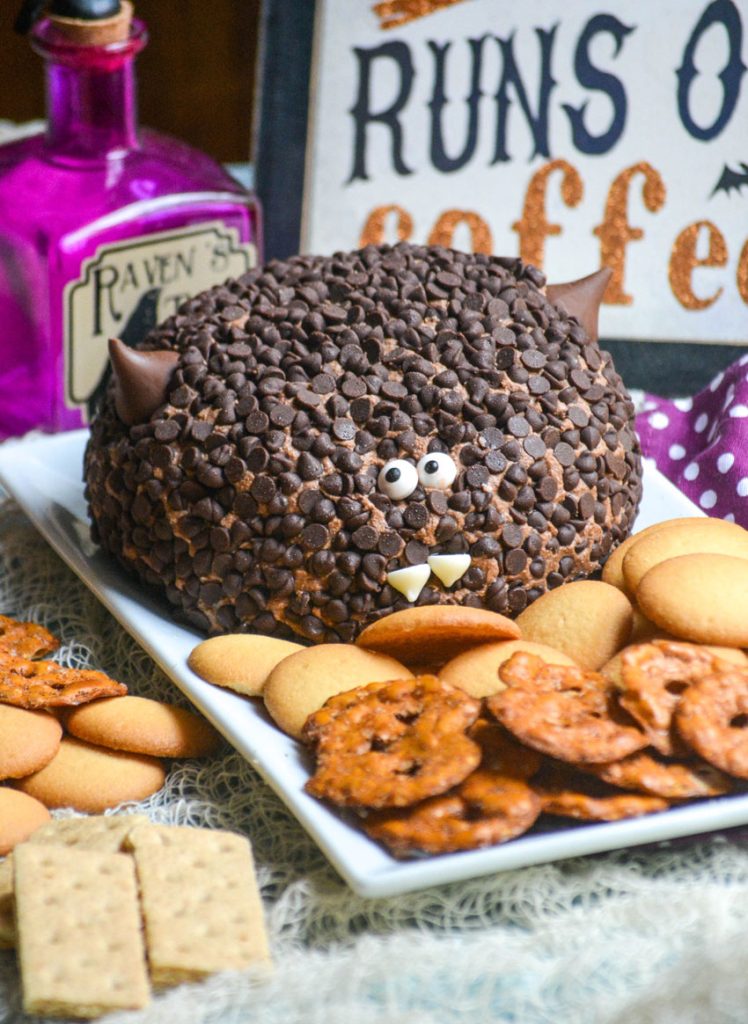 a triple chocolate dessert cheese ball covered in mini chocolate chips and made to look like a bat served on a white platter with pretzel chips and cookies for dipping