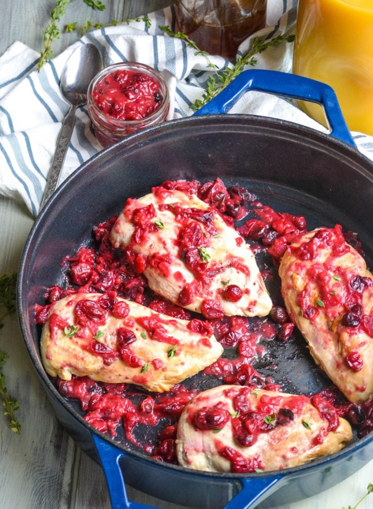 cranberry orange chicken shown in a blue enameled cast iron skillet with extra cranberry sauce and a pitcher of orange juice in the background