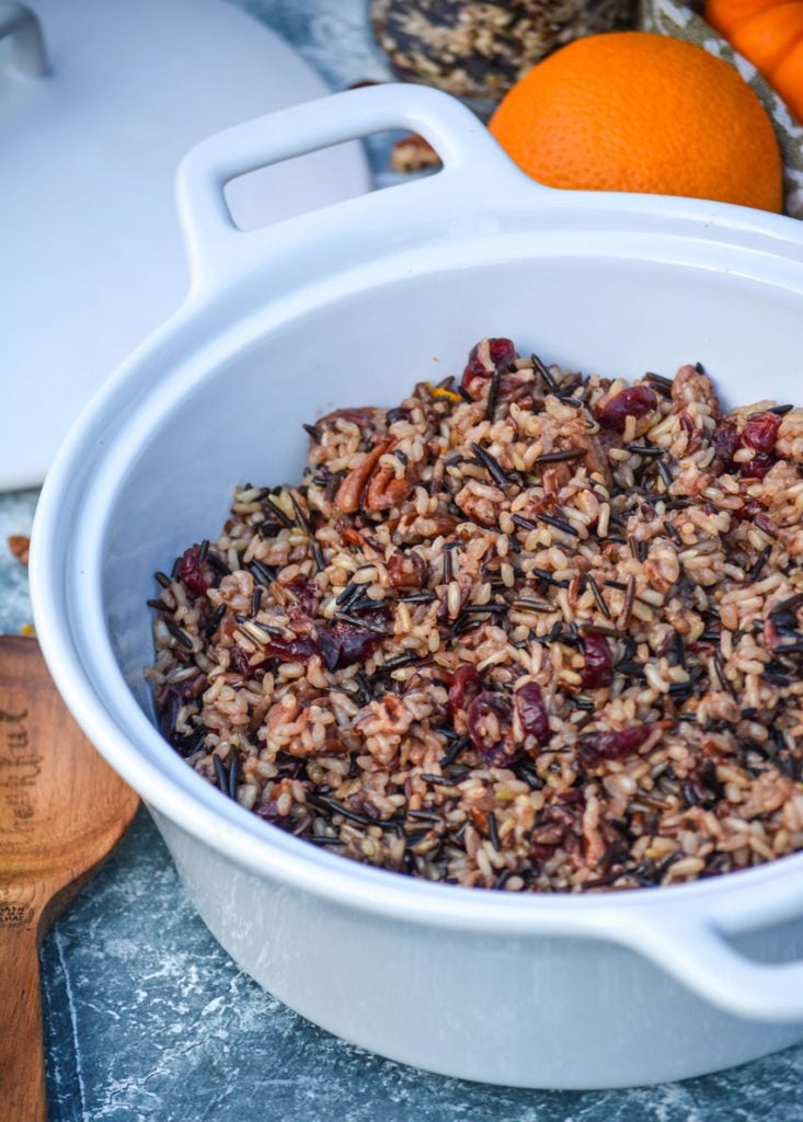cranberry wild rice stuffing shown in double handled white bowl with oranges and a wooden spoon on the sides