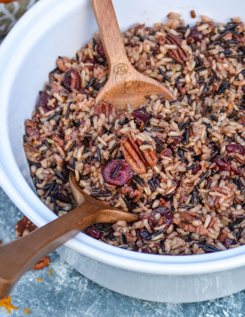 two wooden serving utensils shown scooping cranberry pecan wild rice out of a white serving bowl