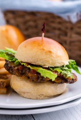 a fully loaded burger using homemade hamburger buns shown on a white plate with a side of fries and bread basket in the background