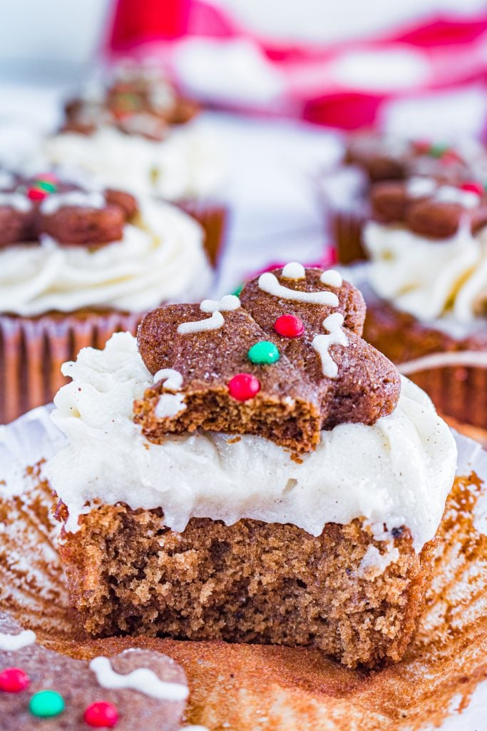 frosted gingerbread cupcakes topped with gingerbread cookies with a bite taken out of the one in focus to show the moist, fluffy cupcake interior
