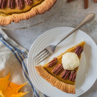 a slice of pumpkin pie with pecans served on a small white plate with silver forks for eating