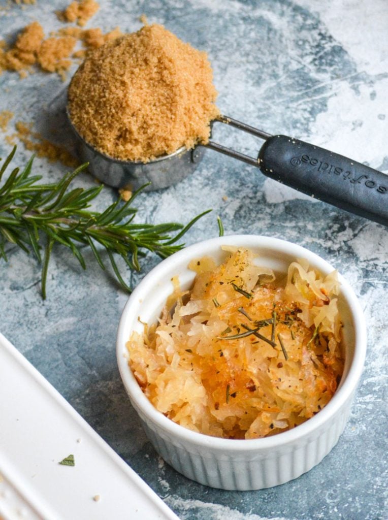 rosemary brown sugar sauerkraut shown in a white ramekin with a measuring cup of brown sugar and a sprig of rosemary in the background