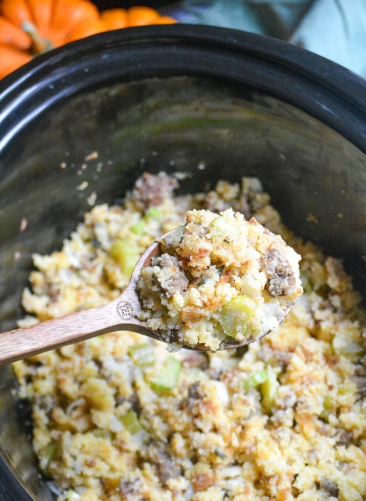 a wooden spoon holding a serving of slow cooker cornbread and sausage stuffing aloft over a crockpot