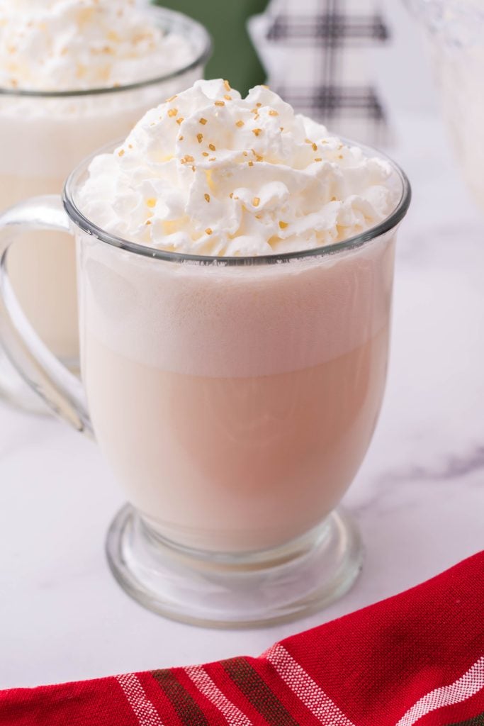 creamy white christmas punch shown in a glass jar, topped with whipped cream and golden sprinkles