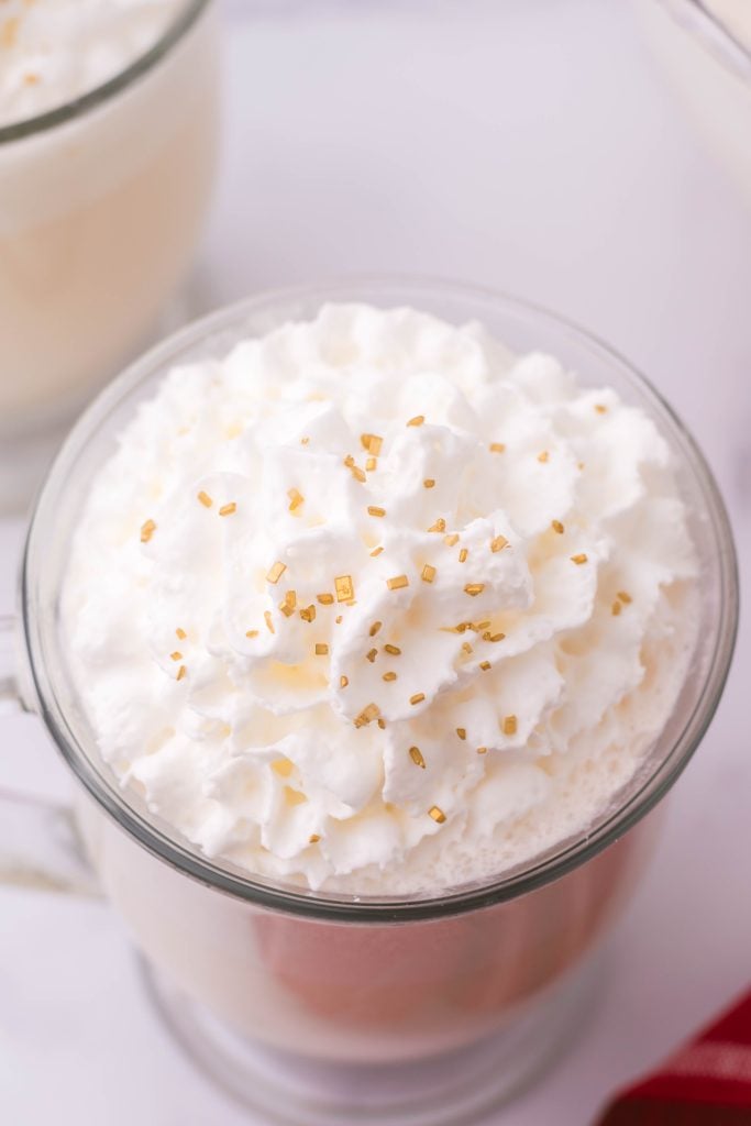 creamy white christmas punch shown in a glass jar, topped with whipped cream and golden sprinkles