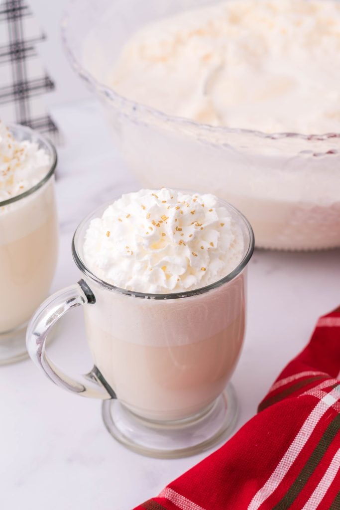 creamy white christmas punch shown in a glass jar, topped with whipped cream and golden sprinkles