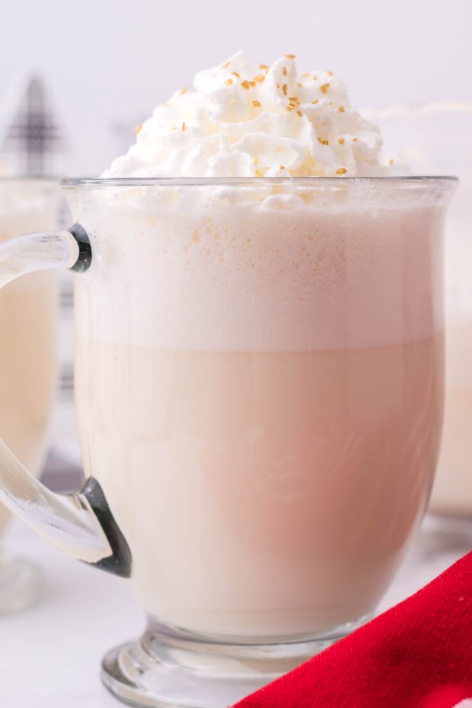 creamy white christmas punch shown in a glass jar, topped with whipped cream and golden sprinkles