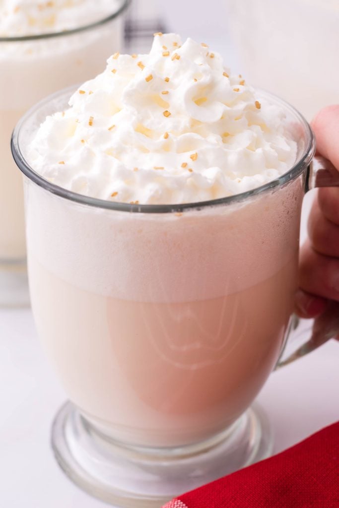 creamy white christmas punch shown in a glass jar, topped with whipped cream and golden sprinkles