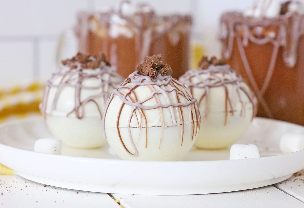 rumchata hot cocoa bombs shown on a white plate with a mug of hot chocolate in the background