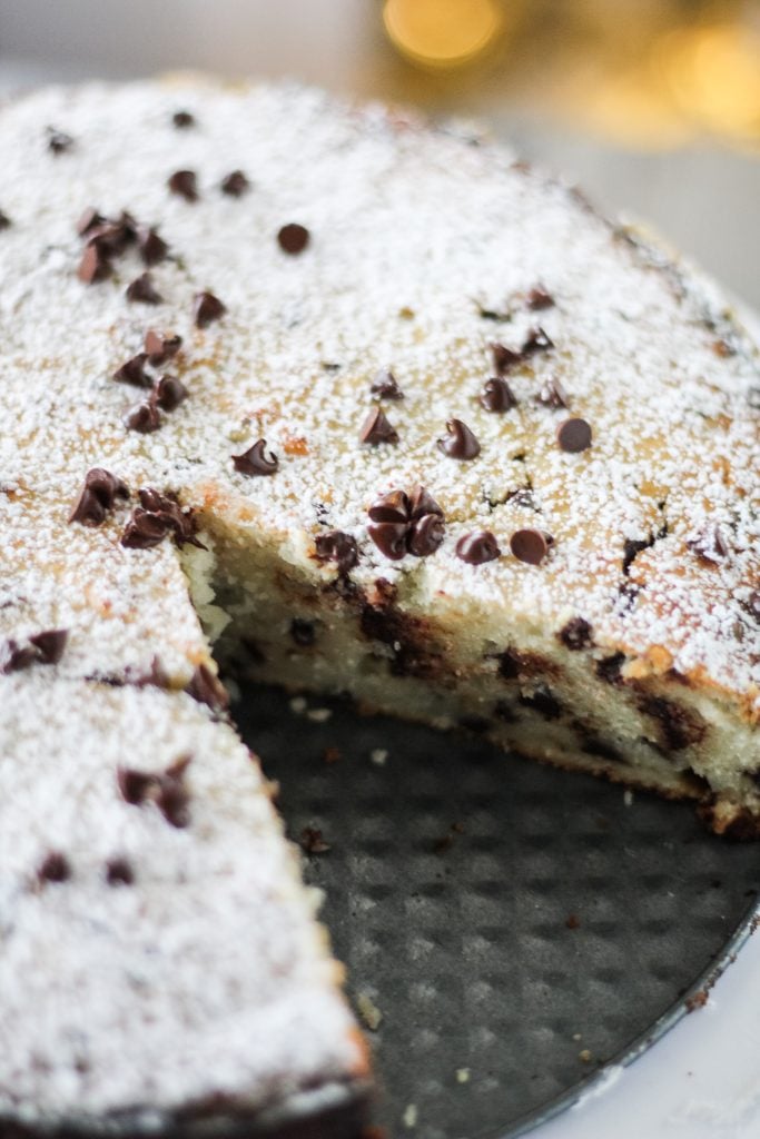 a powdered sugar sprinkled chocolate chip ricotta cake shown on a white cake plate with a slice removed