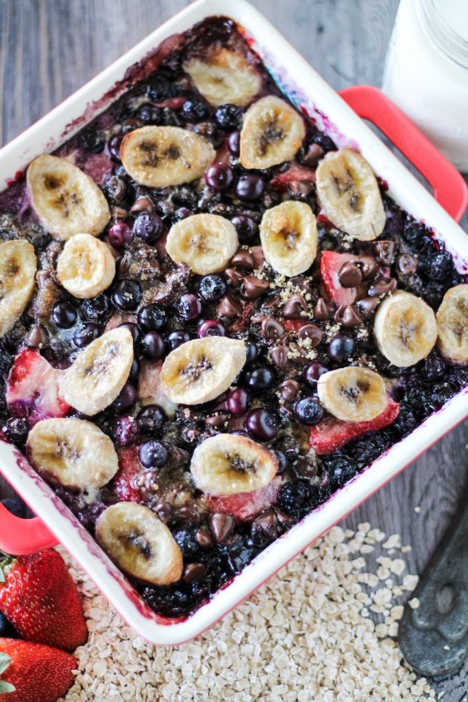 banana berry breakfast bake in a red square baking dish surrounded by fresh berries, oats, and a glass of milk