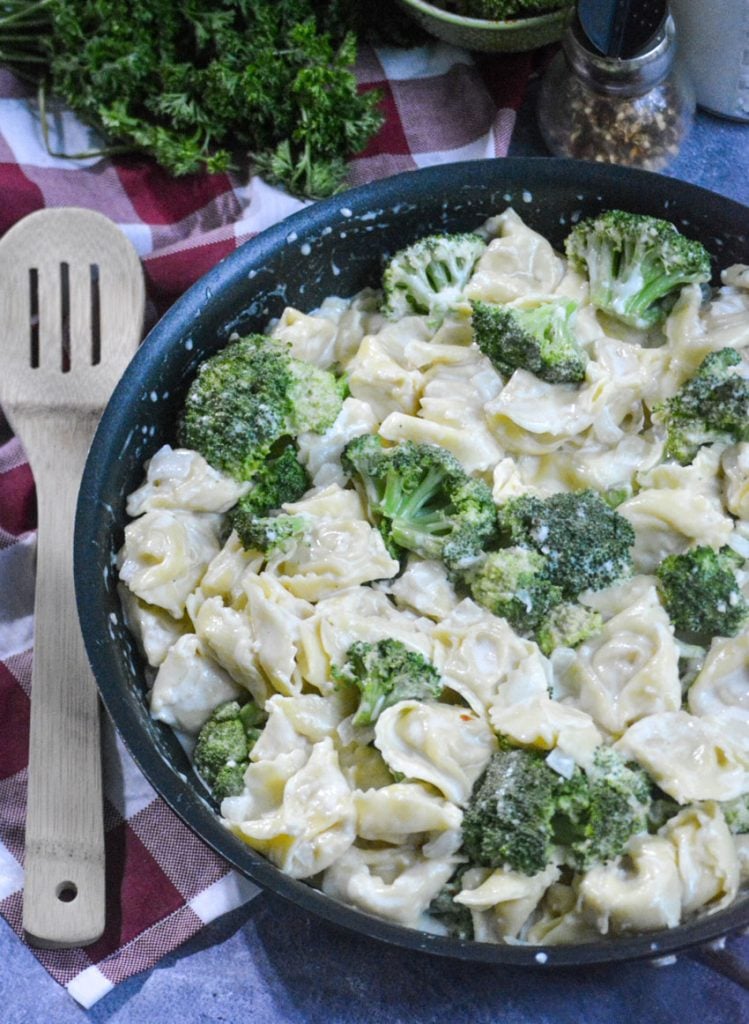 creamy alfredo tortellini with broccoli in a black skillet with a wooden spoon on the side for serving