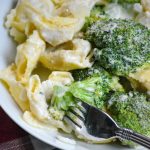 a silver fork digging into creamy alfredo tortellini with broccoli on a white plate