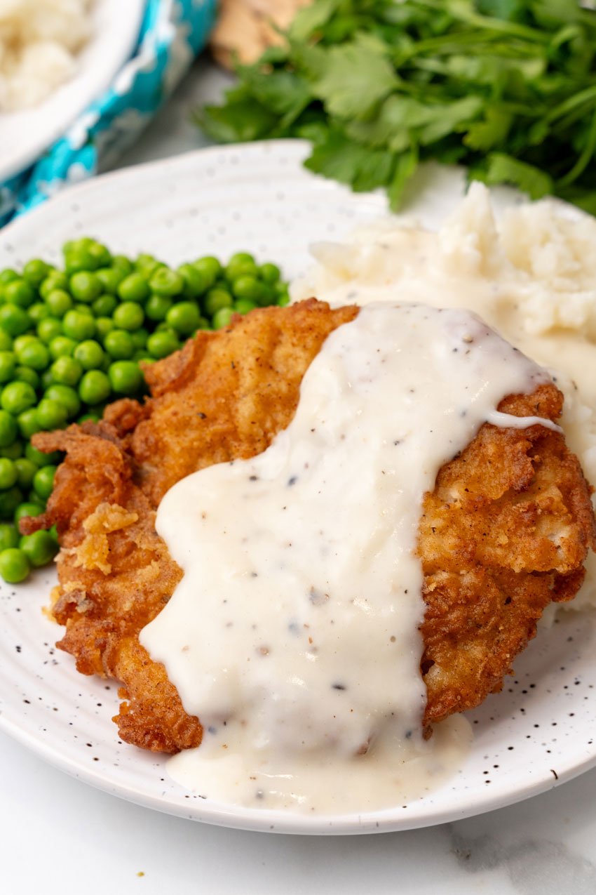 a slice of country fried steak drizzled with white gravy on a white dinner plate with peas and mashed potatoes on the side