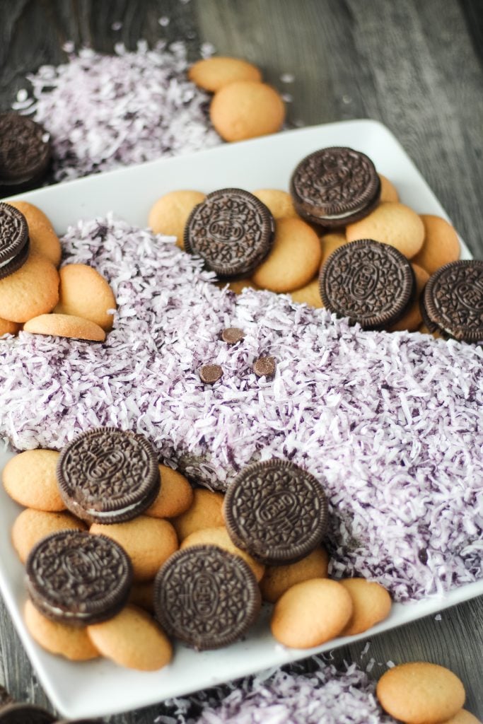 a purple Easter peeps themed cookies and cream cheese ball shown on a white plate surrounded with cookies for dipping