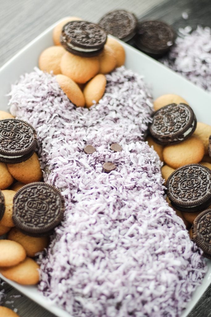 a purple Easter peeps themed cookies and cream cheese ball shown on a white plate surrounded with cookies for dipping