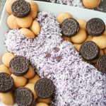 a purple Easter peeps themed cookies and cream cheese ball shown on a white plate surrounded with cookies for dipping