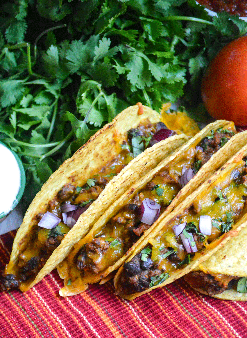 three oven baked tacos lined up with a bunch of fresh cilantro and ripe tomatoes in the background