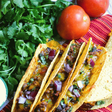 three oven baked tacos lined up with a bunch of fresh cilantro and ripe tomatoes in the background