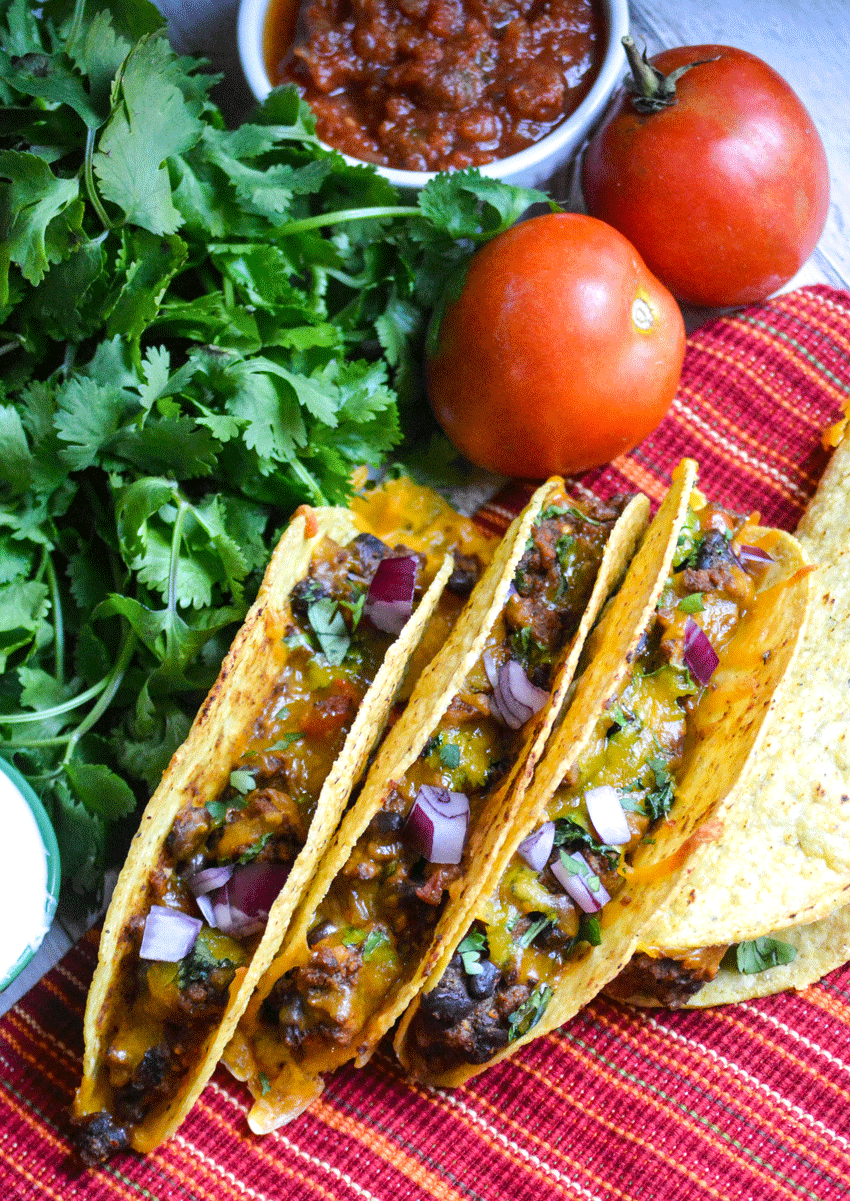 three oven baked tacos lined up with a bunch of fresh cilantro and ripe tomatoes in the background