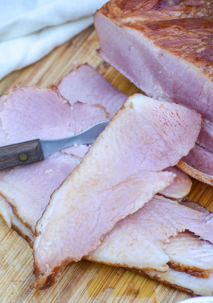 slices of crockpot glazed ham shown on a wooden cutting board with a wooden handled meat fork