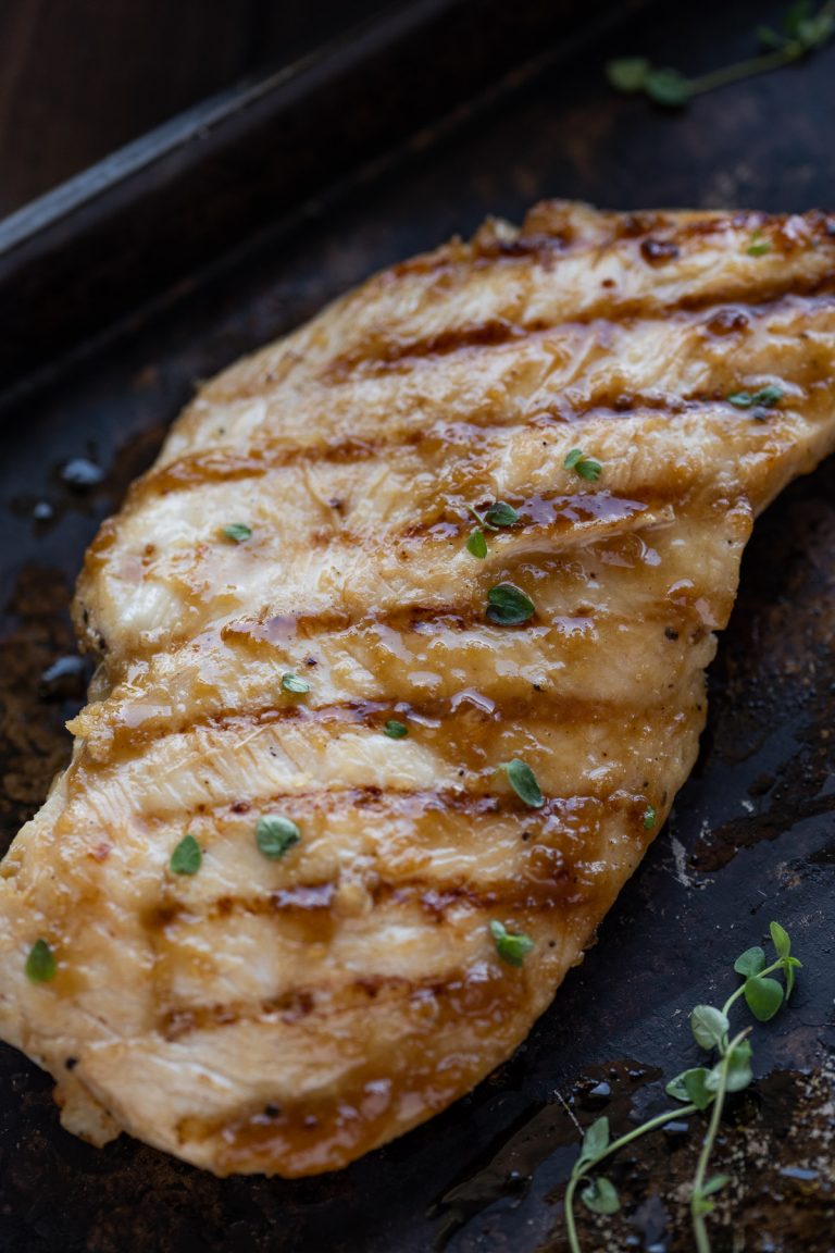 a single grilled teriyaki chicken breast shown on a black sheet pan with fresh herbs