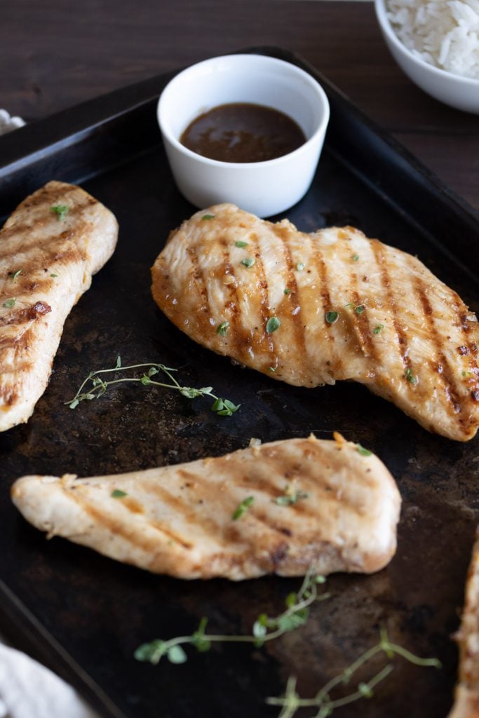 grilled teriyaki chicken on a black sheet pan with fresh herbs and sauce for dipping