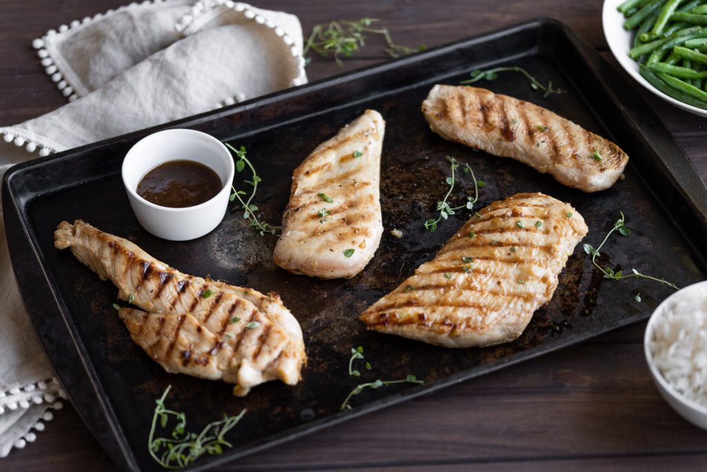 grilled teriyaki chicken on a black sheet pan with fresh herbs and sauce for dipping