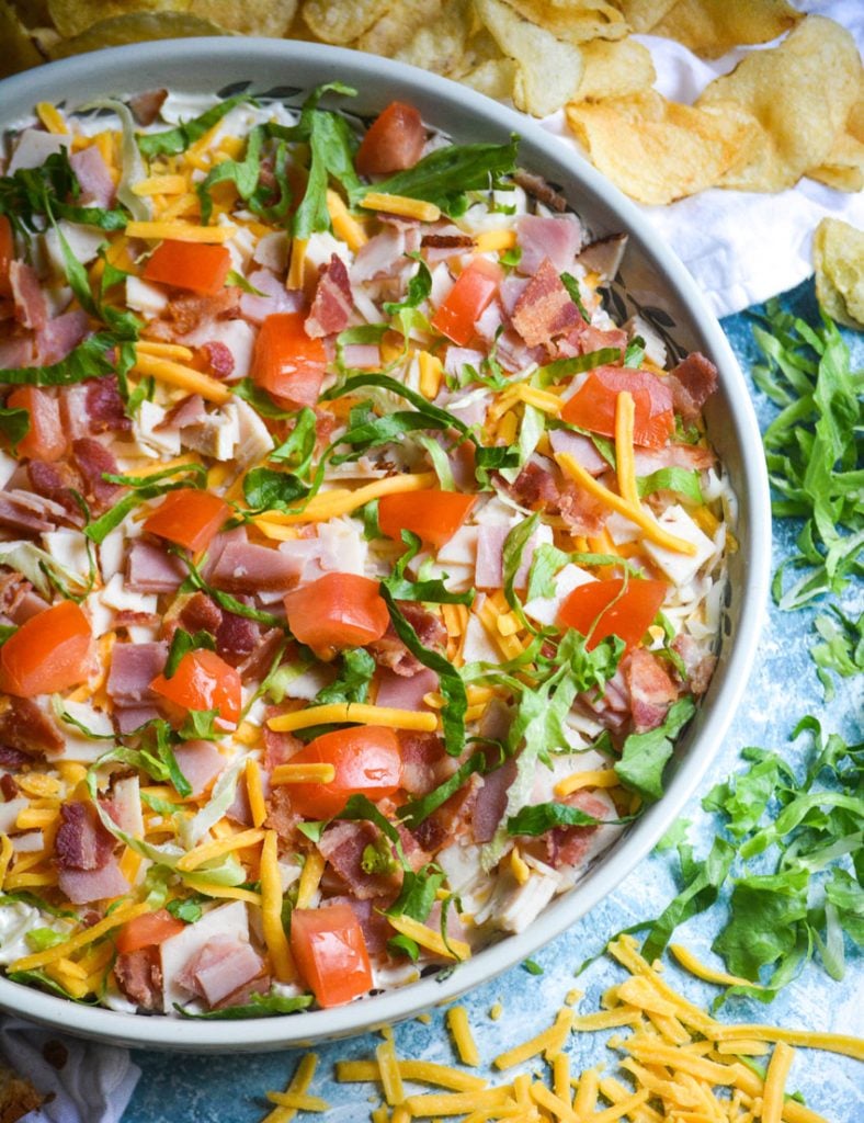 club sandwich dip shown in a large shallow bowl surrounded by chips and triangles of toasts for dipping