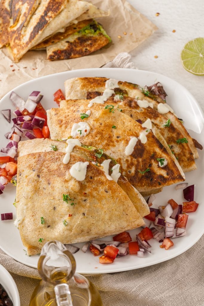 tiktok folded quesadillas stacked on a white plate, topped with tomatoes, red onions, cilantro, and drizzled with sour cream