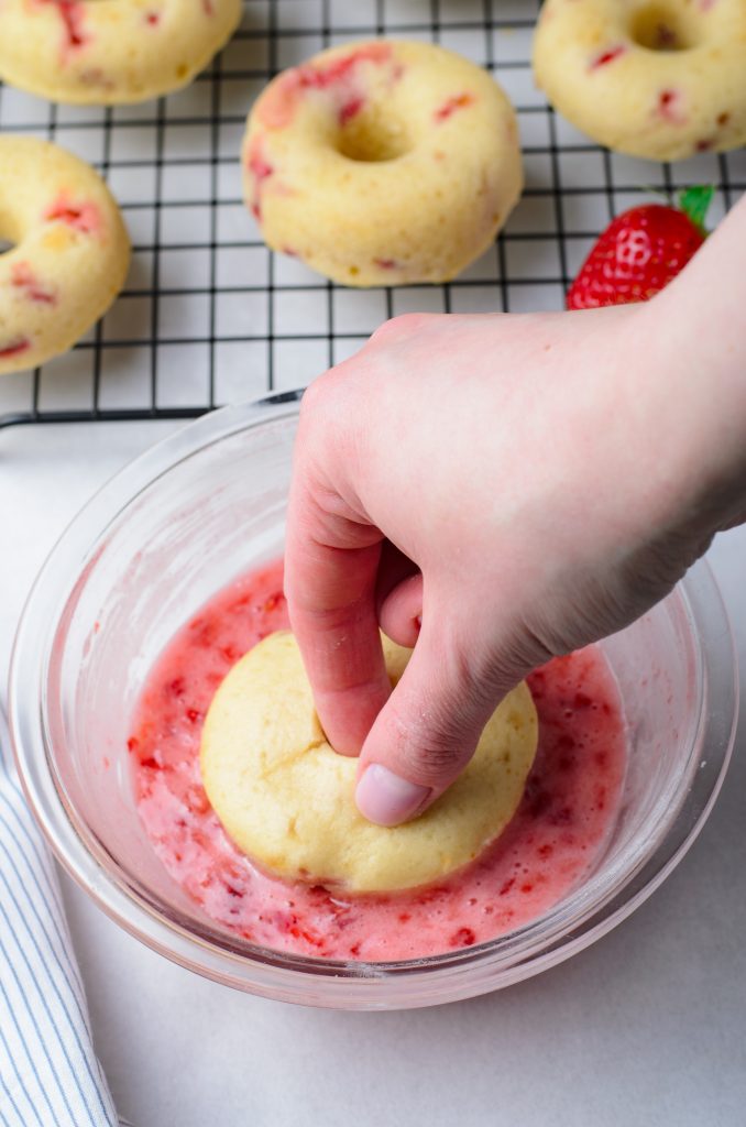 a hand shown dunking a strawberry donut into a bowl filled with pink glaze