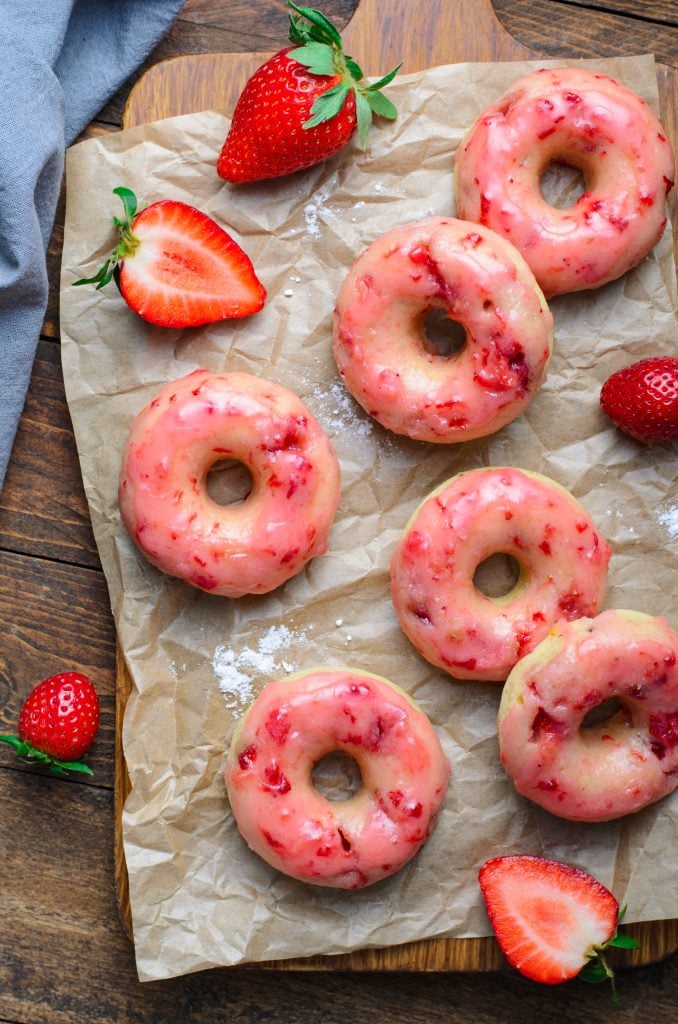 a batch of baked strawberry glazed donuts shown on crinkled brown parchment paper with fresh berries in the background